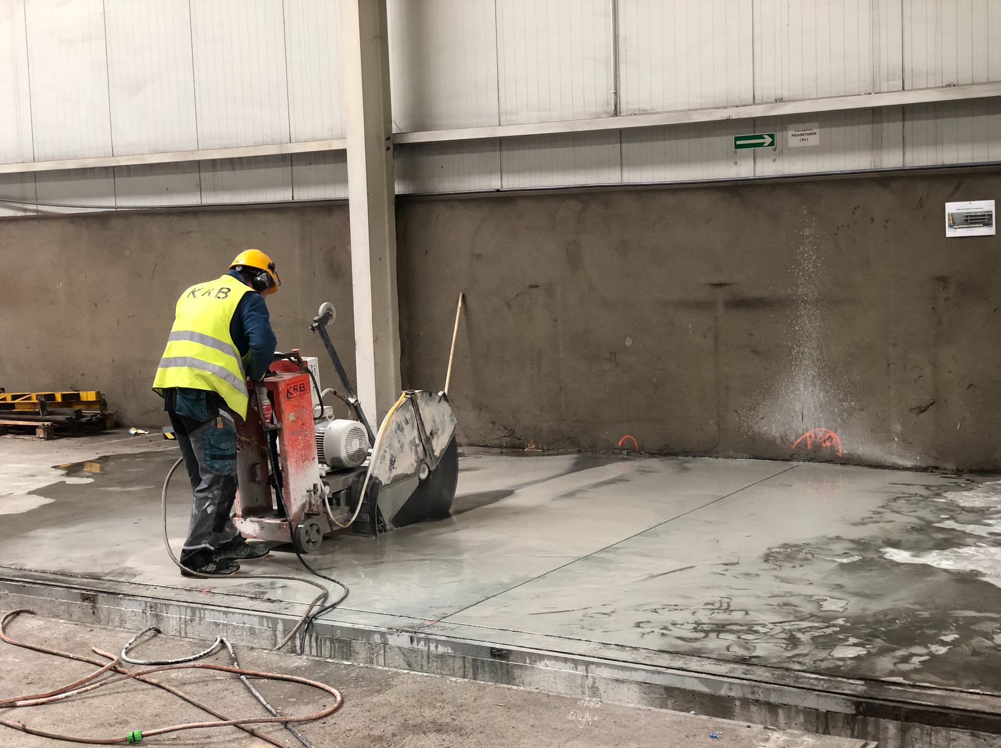 Pracownik w żółtej kamizelce tnie betonową posadzkę przecinarką z tarczą diamentową w hali przemysłowej. Widoczne linie cięcia i rozpryskująca woda.