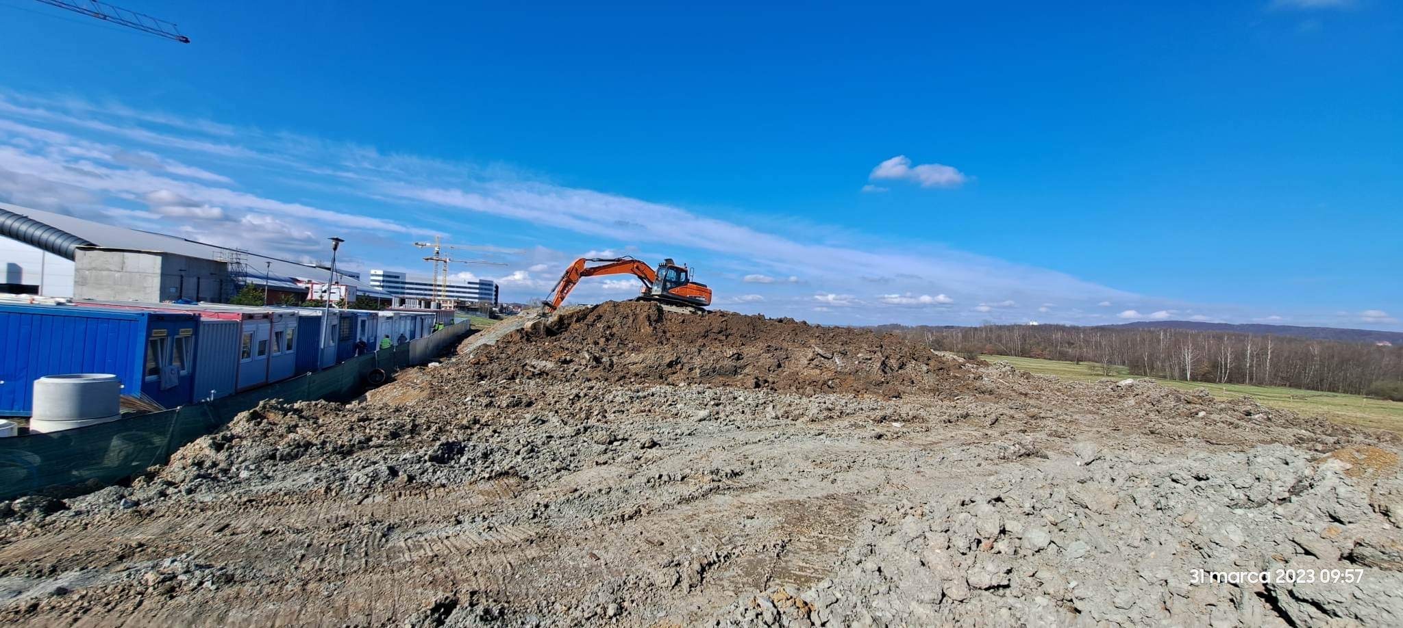 Pomarańczowa koparka na wzgórzu ziemi, kontenery budowlane w tle, błękitne niebo z chmurami. Prace ziemne w toku, widok na budynki w oddali.