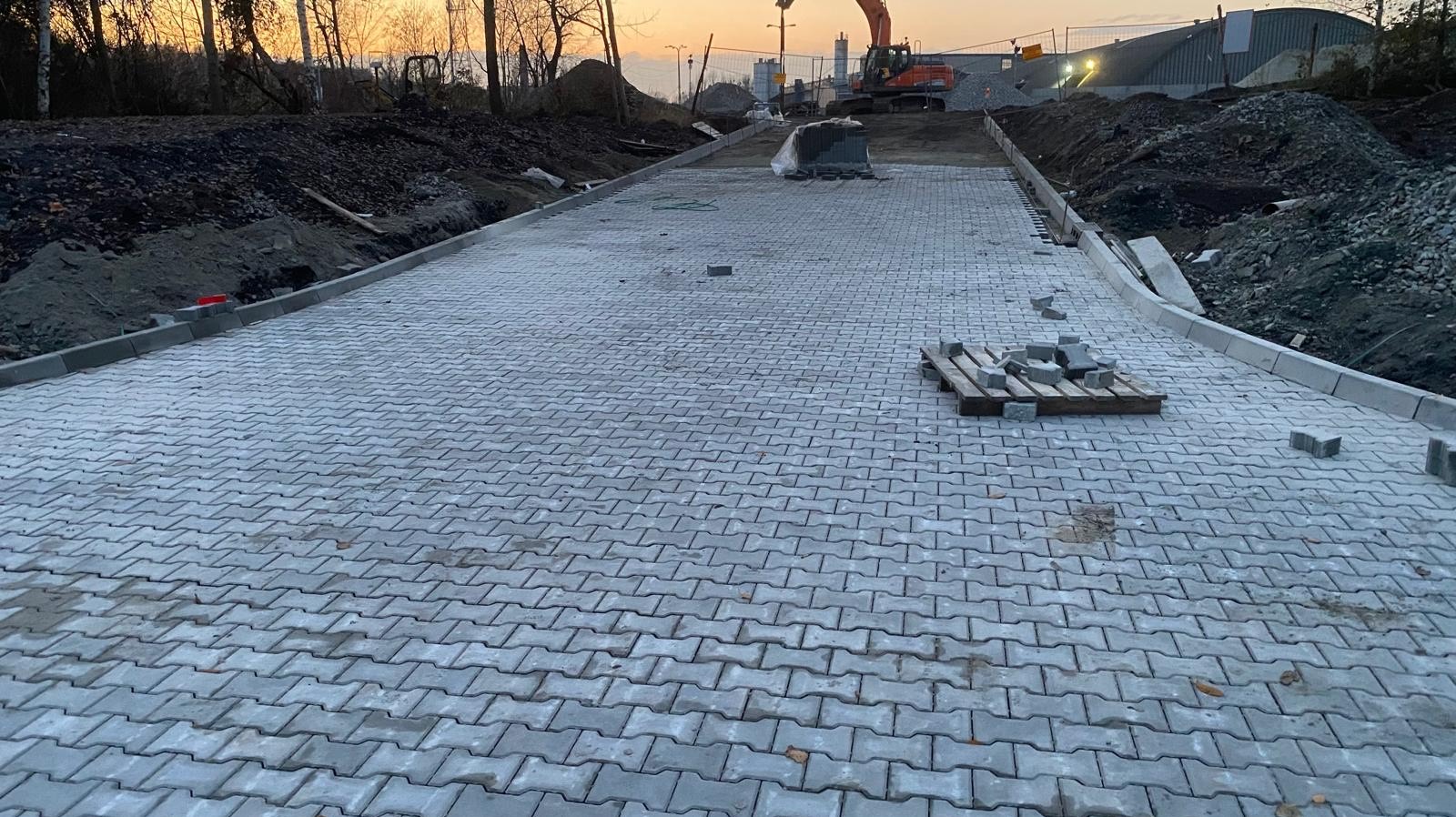 Układanie szarej kostki brukowej na nowym chodniku, widoczny paleta z kostką, krawężniki i koparka w tle, oświetlone zachodzącym słońcem.