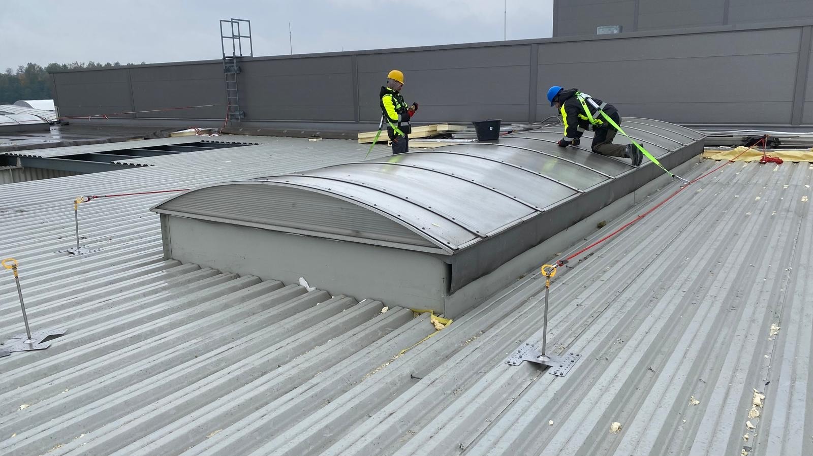 Dwóch pracowników w uprzężach na dachu z blachy trapezowej, montujących świetlik dachowy. Widoczne zabezpieczenia linowe i narzędzia pracy.