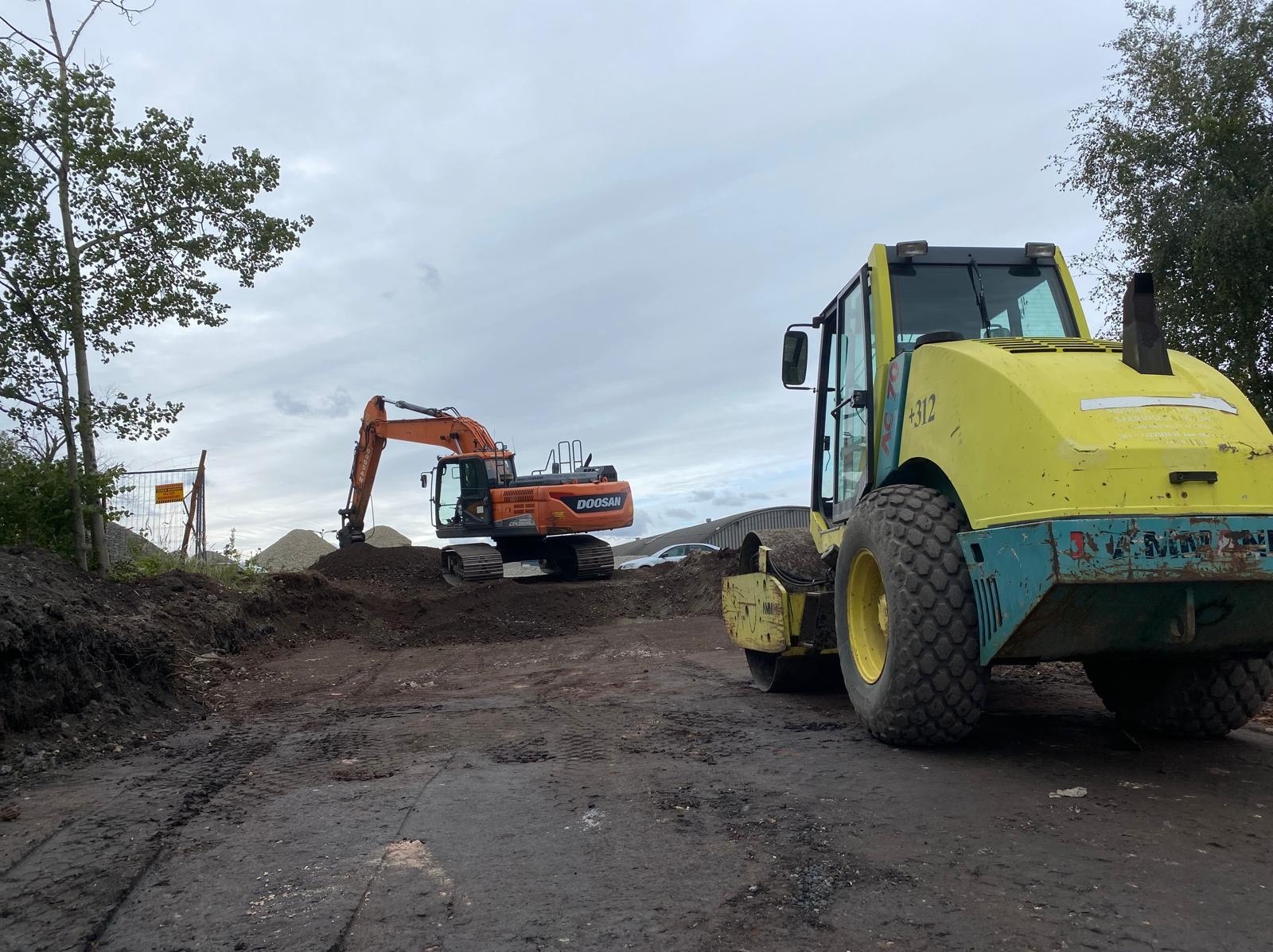 Pomarańczowa koparka Doosan i żółty walec na placu budowy z ziemią i gruzem, pod pochmurnym niebem. Widoczne ślady opon na ubitej ziemi.