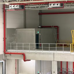 Joshua Drojetzki Media - Instalacja wentylacyjna z czerwonymi rurami w hali magazynowej. Widoczne białe kratki wentylacyjne pod sufitem i drabina oparta o ścianę. Ujęcie z dołu.