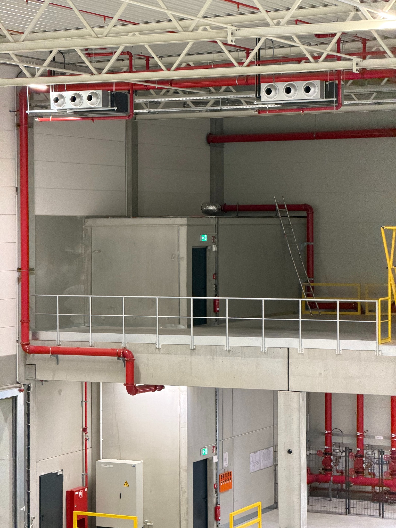 Instalacja wentylacyjna z czerwonymi rurami w hali magazynowej. Widoczne białe kratki wentylacyjne pod sufitem i drabina oparta o ścianę. Ujęcie z dołu.