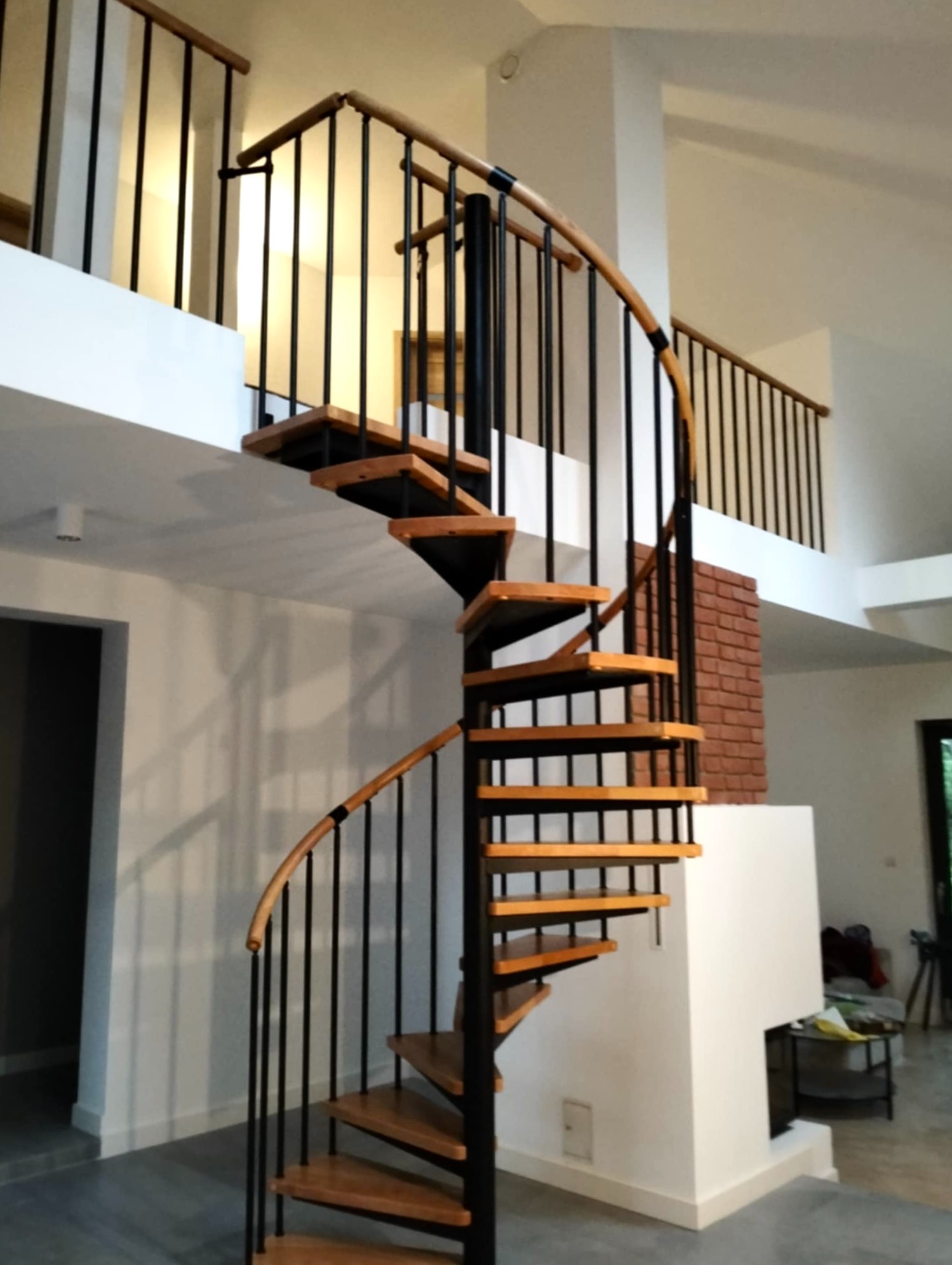Spiralne schody z drewnianymi stopniami i czarną metalową konstrukcją w jasnym wnętrzu z antresolą i ceglanym elementem dekoracyjnym.