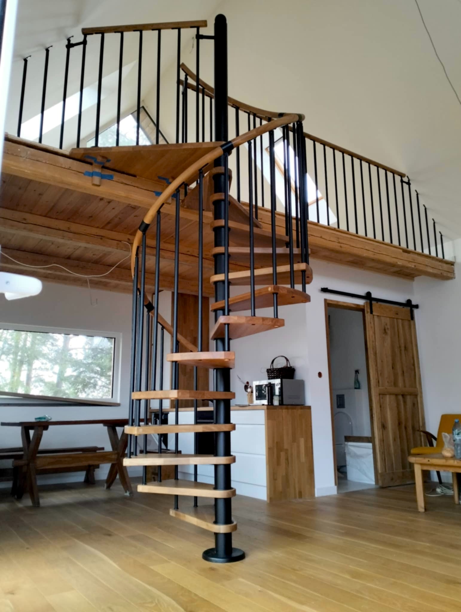 Spiralne schody z drewnianymi stopniami i czarną metalową konstrukcją w przestronnym wnętrzu domu w Jerce. Widoczna balustrada na antresoli.