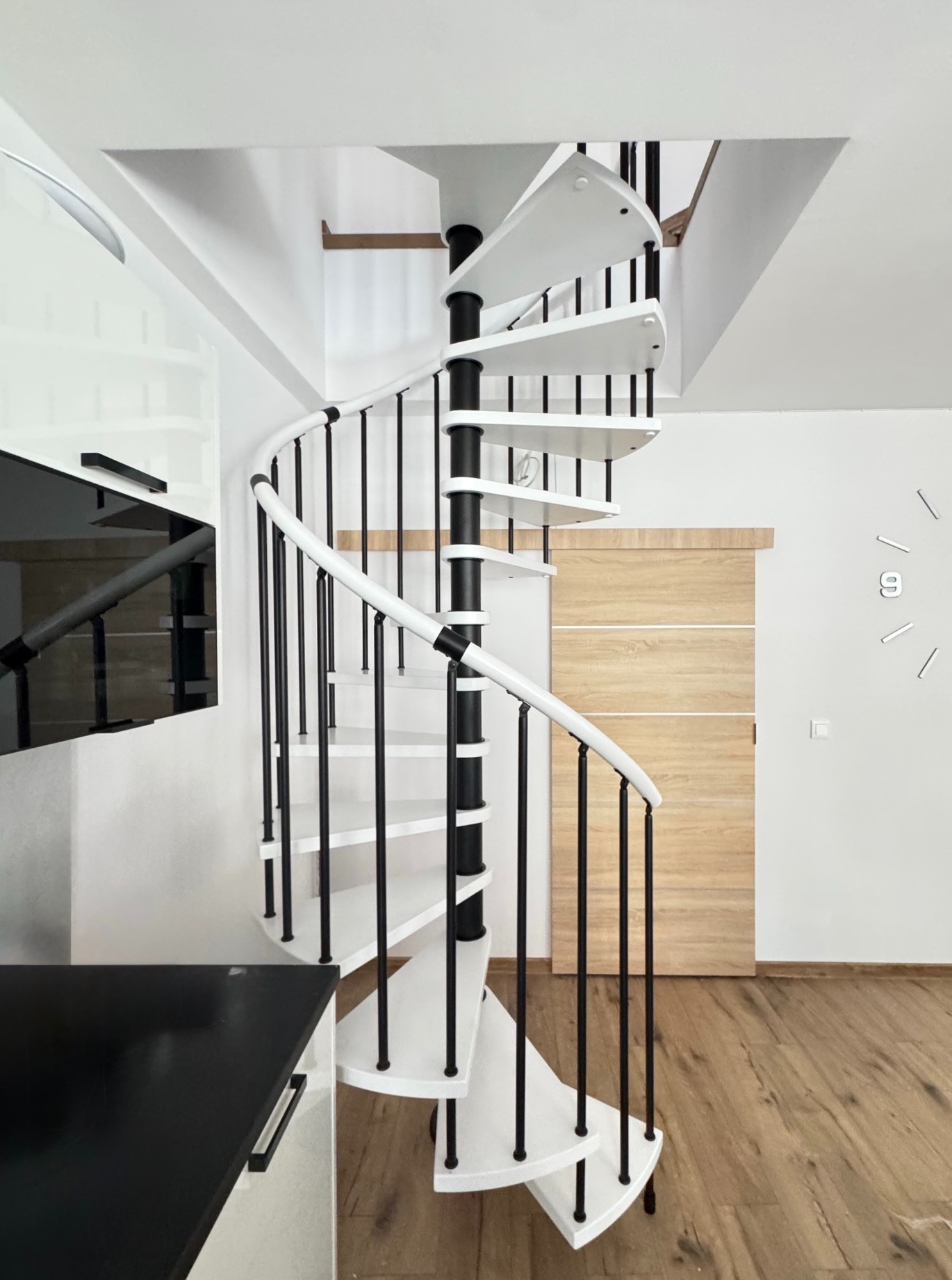 Białe schody spiralne z czarnymi elementami w nowoczesnym wnętrzu. Widoczne drewniane panele podłogowe i minimalistyczny zegar na ścianie.