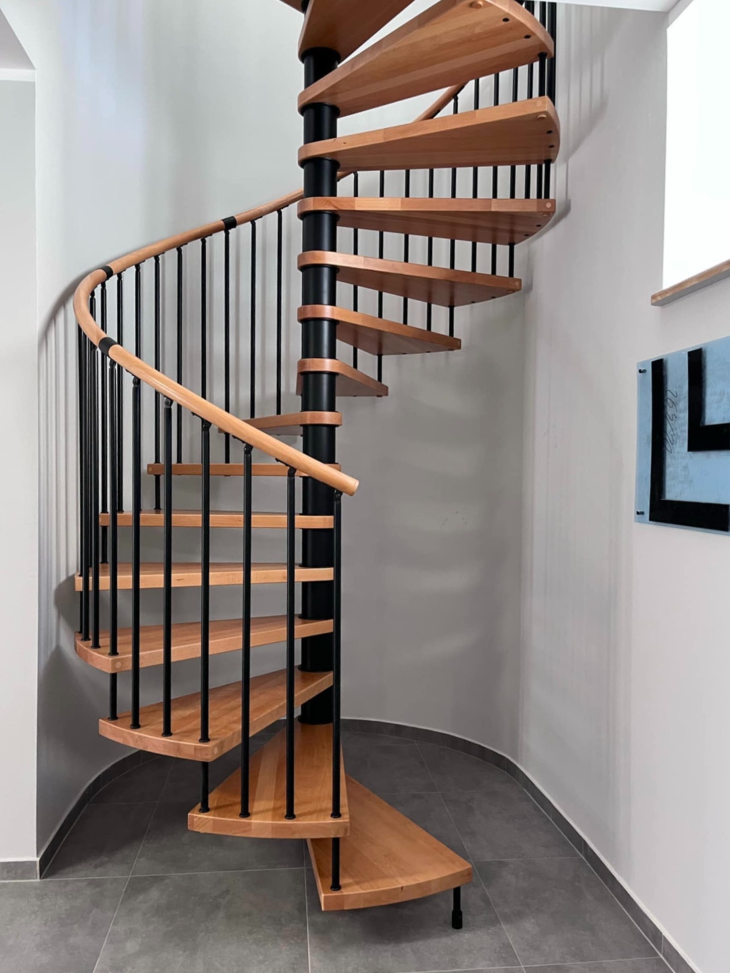 Spiralne schody z drewna bukowego z czarną, metalową konstrukcją i jasną poręczą, umieszczone w rogu pomieszczenia z szarą podłogą i minimalistycznym wystrojem.