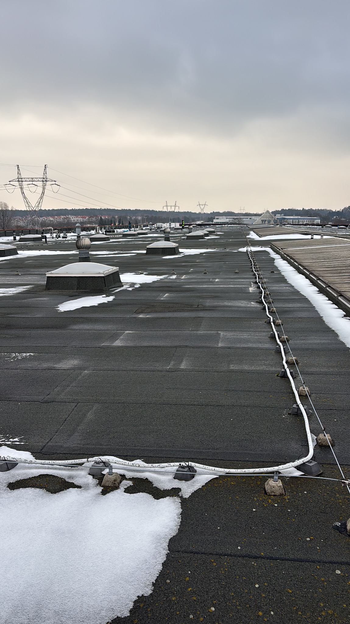 Płaski dach pokryty papą z świetlikami i instalacją odgromową, częściowo pokryty śniegiem. W tle słupy wysokiego napięcia i zabudowania.