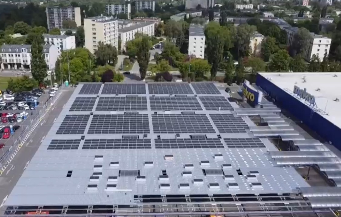 Widok z góry na dużą instalację fotowoltaiczną na dachu budynku Makro w Łodzi. Panele ułożone w rzędach, widoczne otoczenie miejskie i parking.