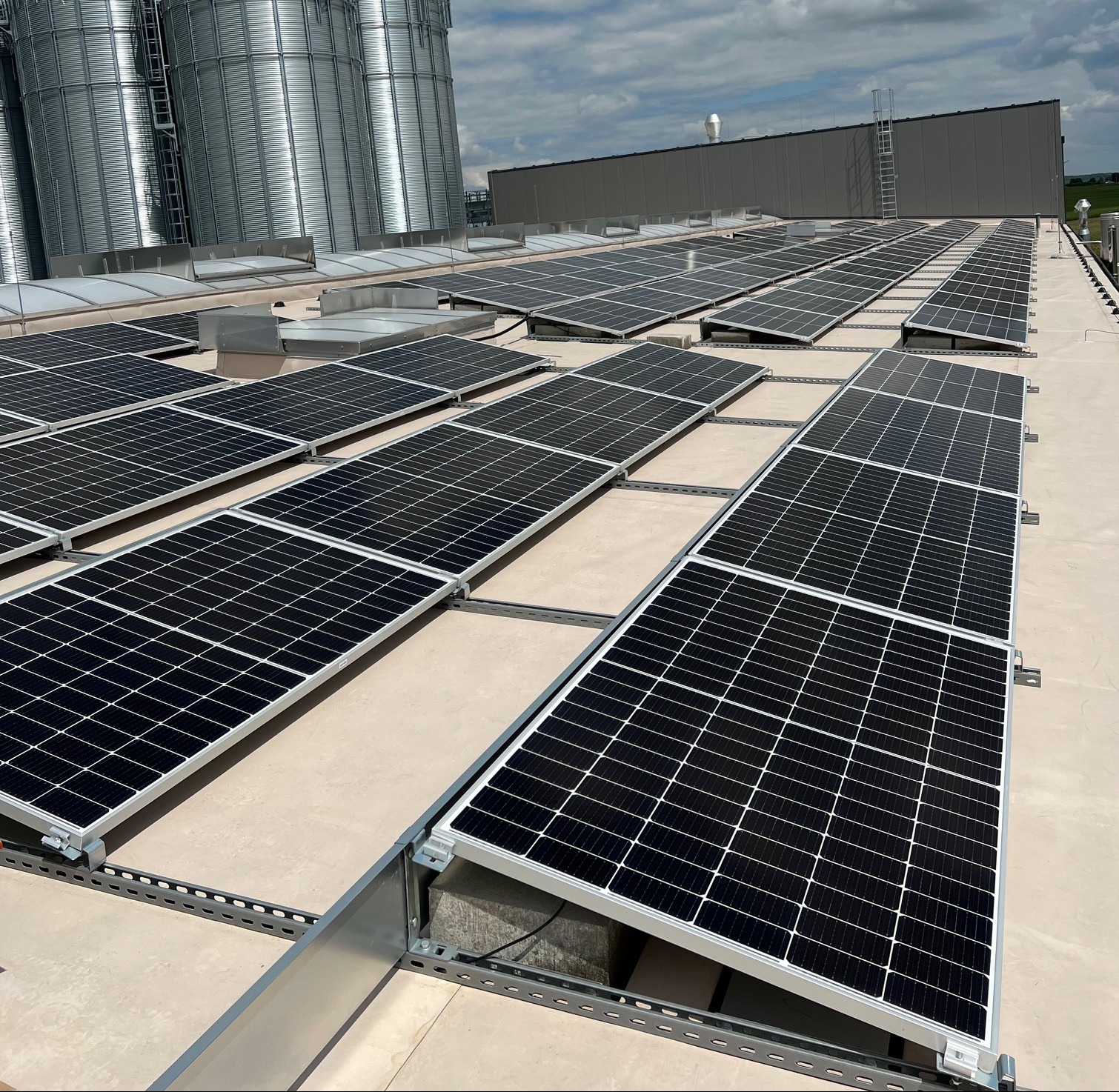 Rozległa instalacja fotowoltaiczna na dachu budynku przemysłowego, z widocznymi silosami w tle i pochmurnym niebem. Panele ułożone w rzędach, metalowe konstrukcje montażowe.