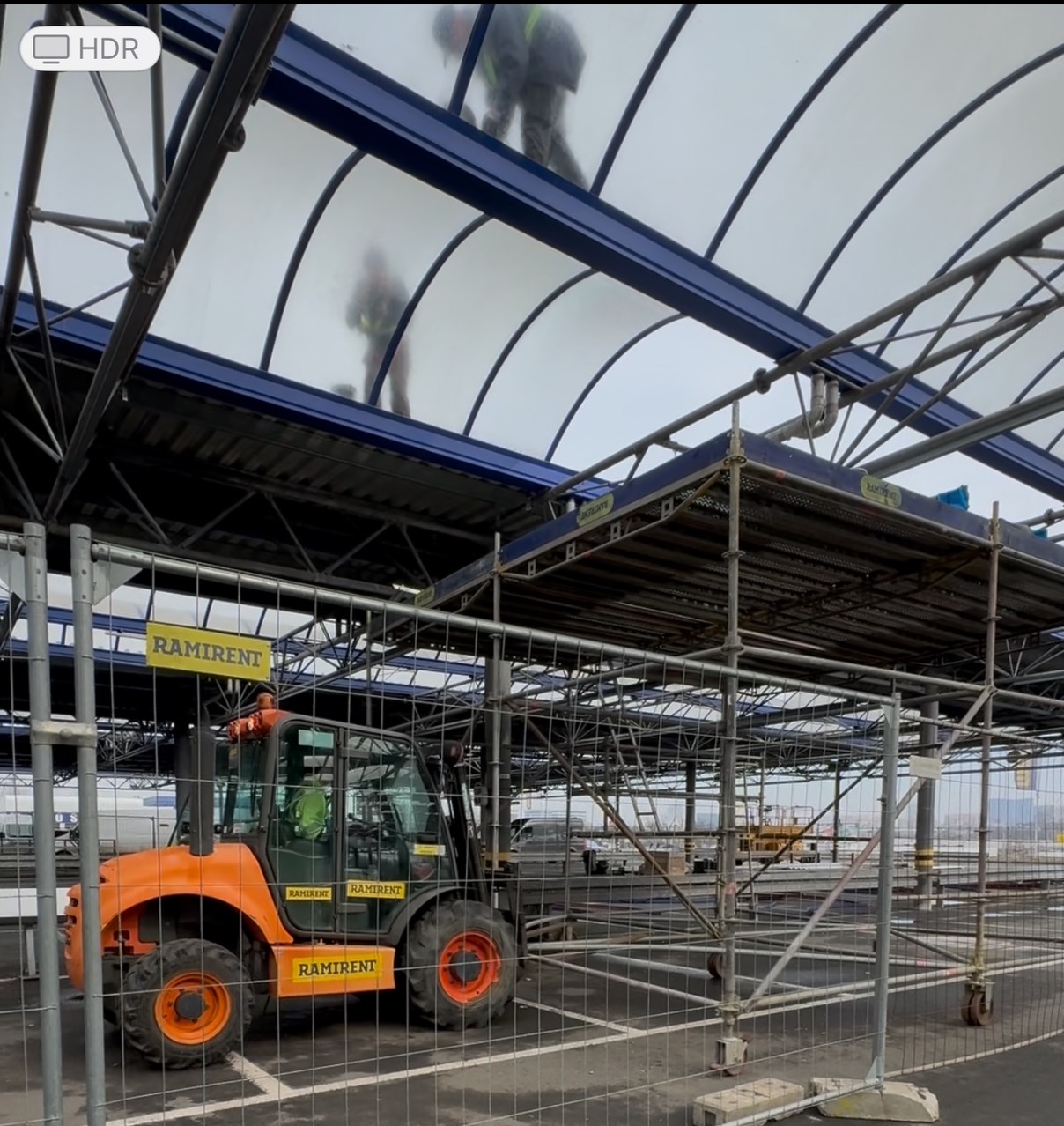 Montaż zadaszenia parkingu w Poznaniu. Widoczne rusztowania, podnośnik Ramirent i pracownicy na dachu. Konstrukcja stalowa z przezroczystymi panelami.