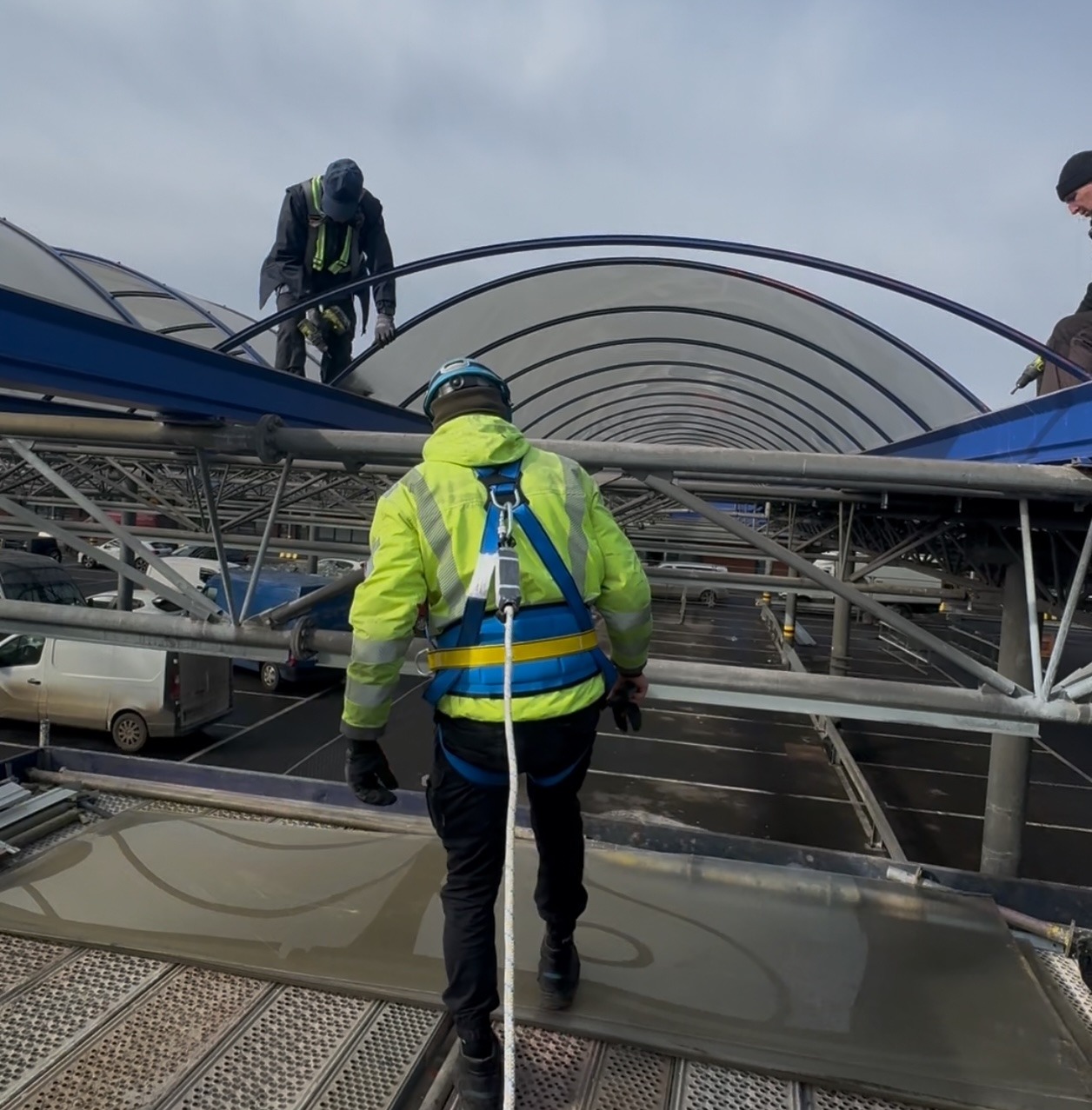 Pracownicy w uprzężach montują zadaszenie nad parkingiem. Widoczne stalowe konstrukcje i częściowo samochody w tle. Jeden z pracowników używa wkrętarki.