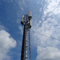 Biuro Projektowe A&A - Metalowa wieża telekomunikacyjna z antenami na tle błękitnego nieba z chmurami. Widok z dołu, ukazujący konstrukcję i elementy mocujące.