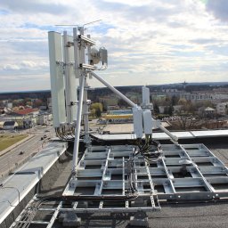 Biuro Projektowe A&A - Anteny telekomunikacyjne zamontowane na dachu budynku z widokiem na miasto w tle. Metalowa konstrukcja wsporcza, kable i urządzenia. Słoneczny dzień.