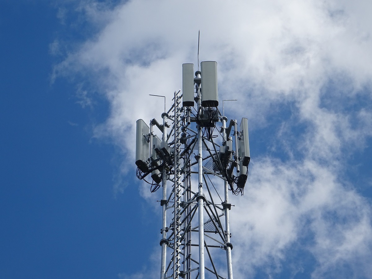 Metalowa wieża telekomunikacyjna z antenami na tle błękitnego nieba z białymi obłokami, widziana z dołu. Konstrukcja kratowa, symetryczny układ anten.