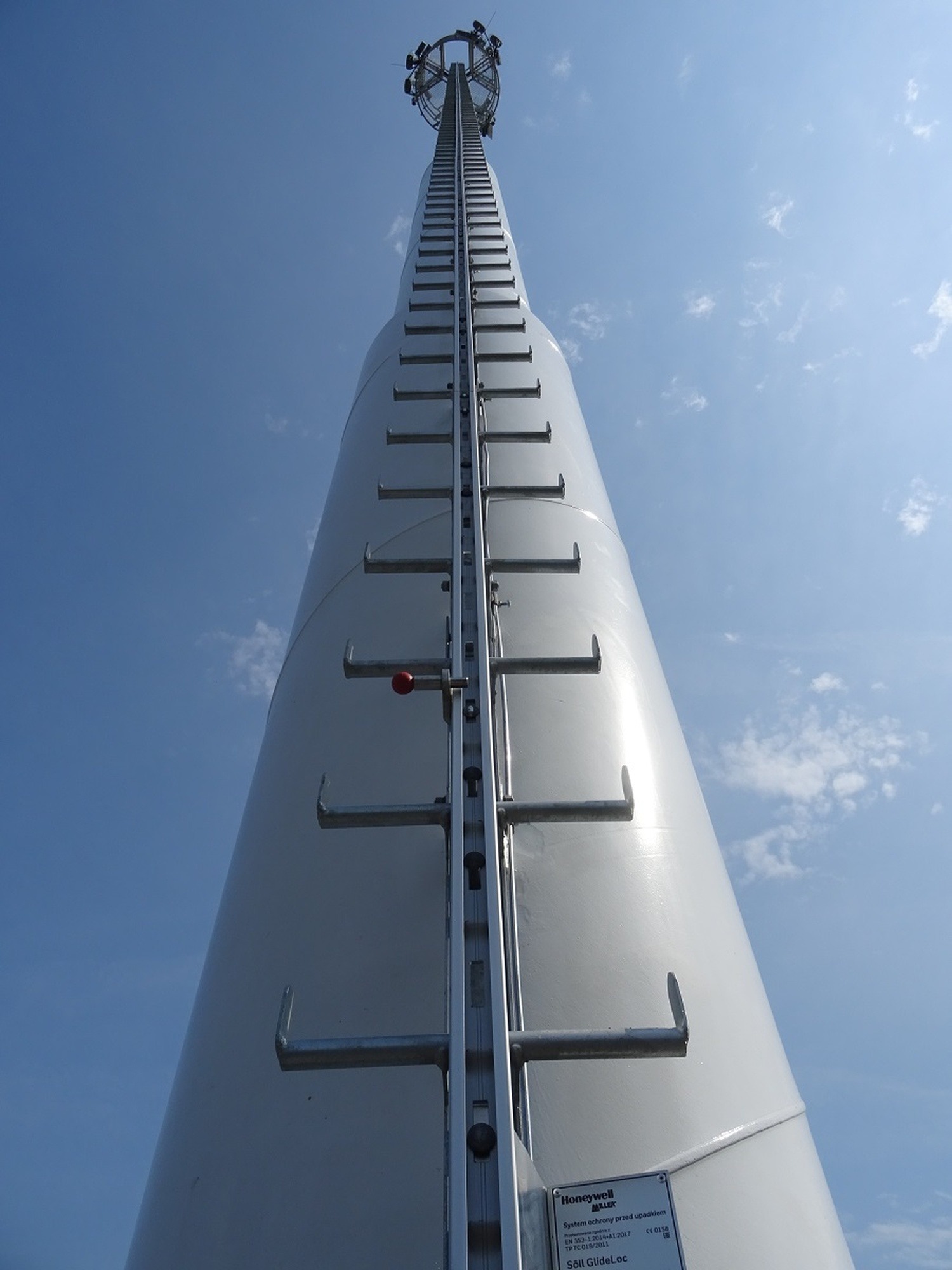Wysoka, biała wieża z drabiną na tle błękitnego nieba. Widok z dołu, podkreślający jej wysokość i konstrukcję. Widoczny system zabezpieczeń przed upadkiem.