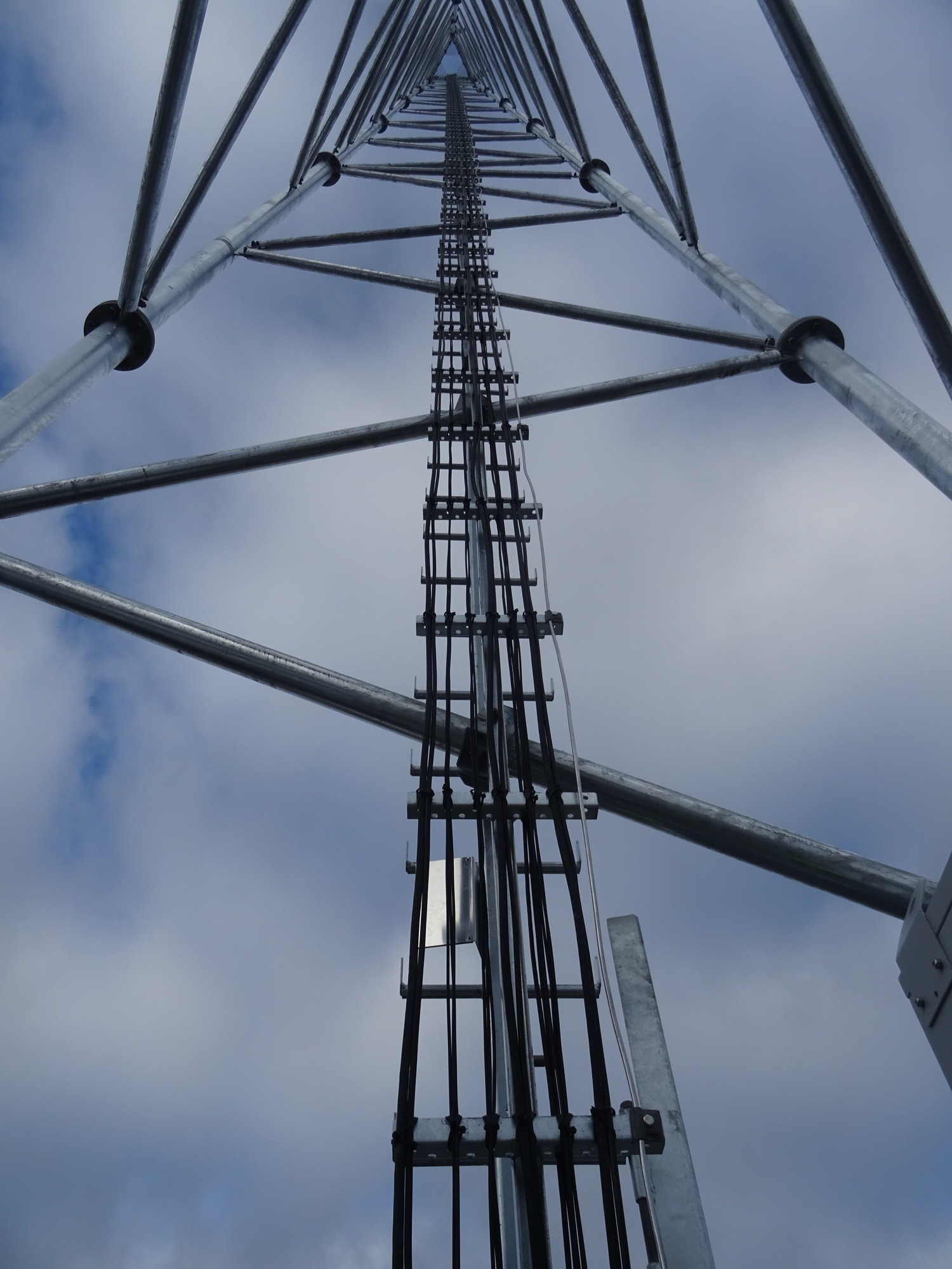 Metalowa konstrukcja wieży telekomunikacyjnej z widocznymi kablami i elementami mocującymi, sfotografowana z dołu na tle pochmurnego nieba.