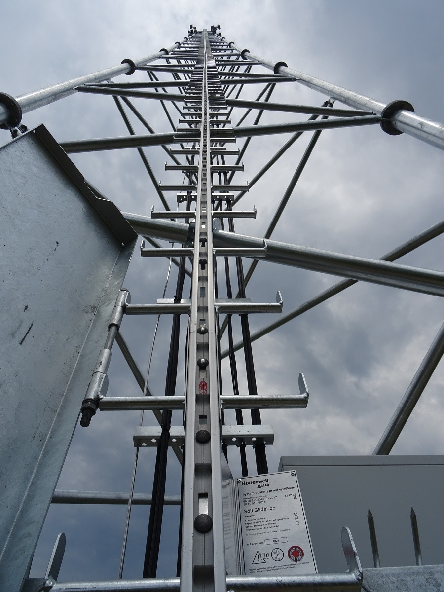 Metalowa konstrukcja wieży telekomunikacyjnej z systemem asekuracyjnym Söll GlideLoc, widok z dołu na tle pochmurnego nieba.