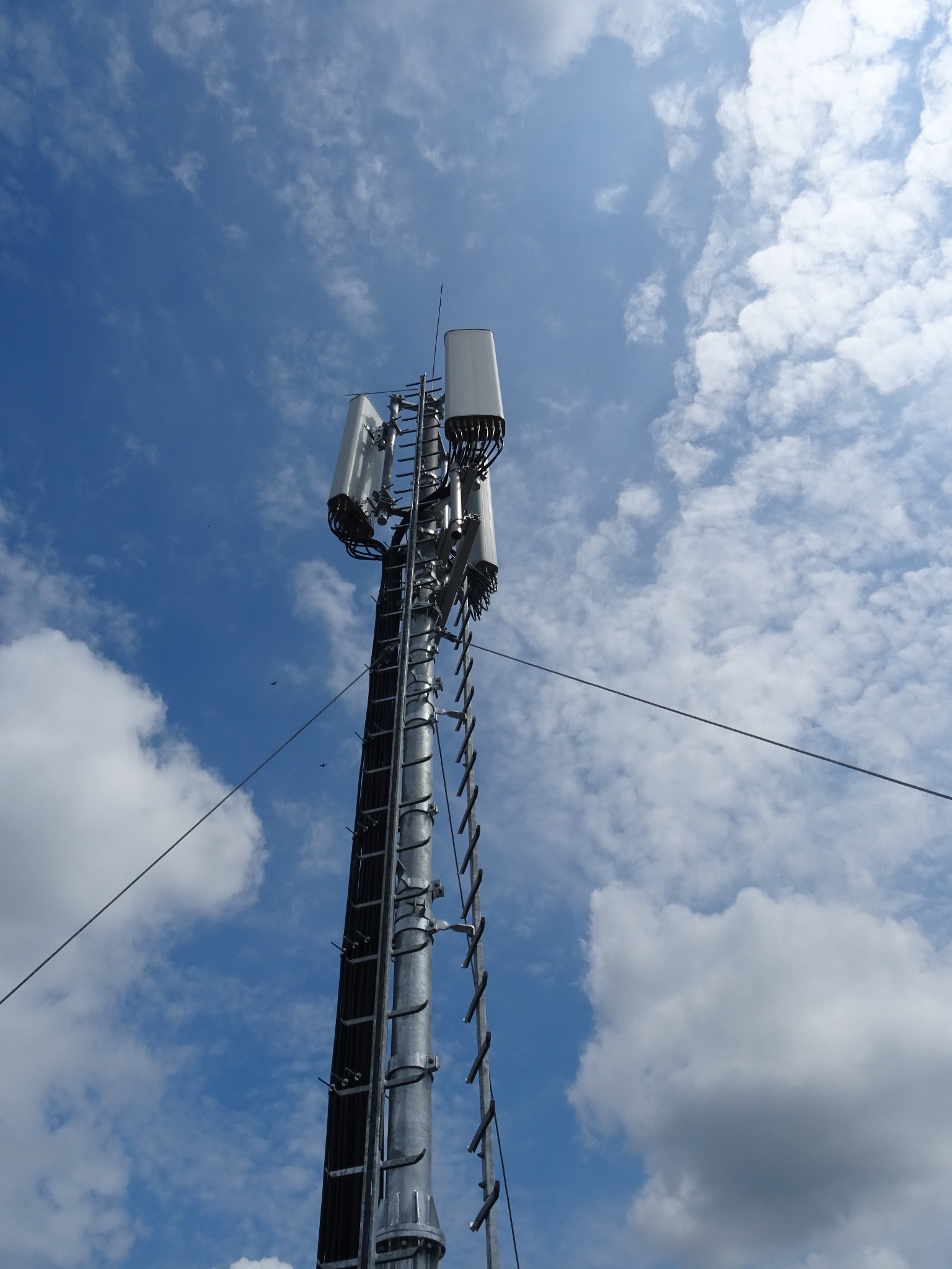 Metalowa wieża telekomunikacyjna z antenami na tle błękitnego nieba z chmurami. Widok z dołu, ukazujący konstrukcję i elementy mocujące.