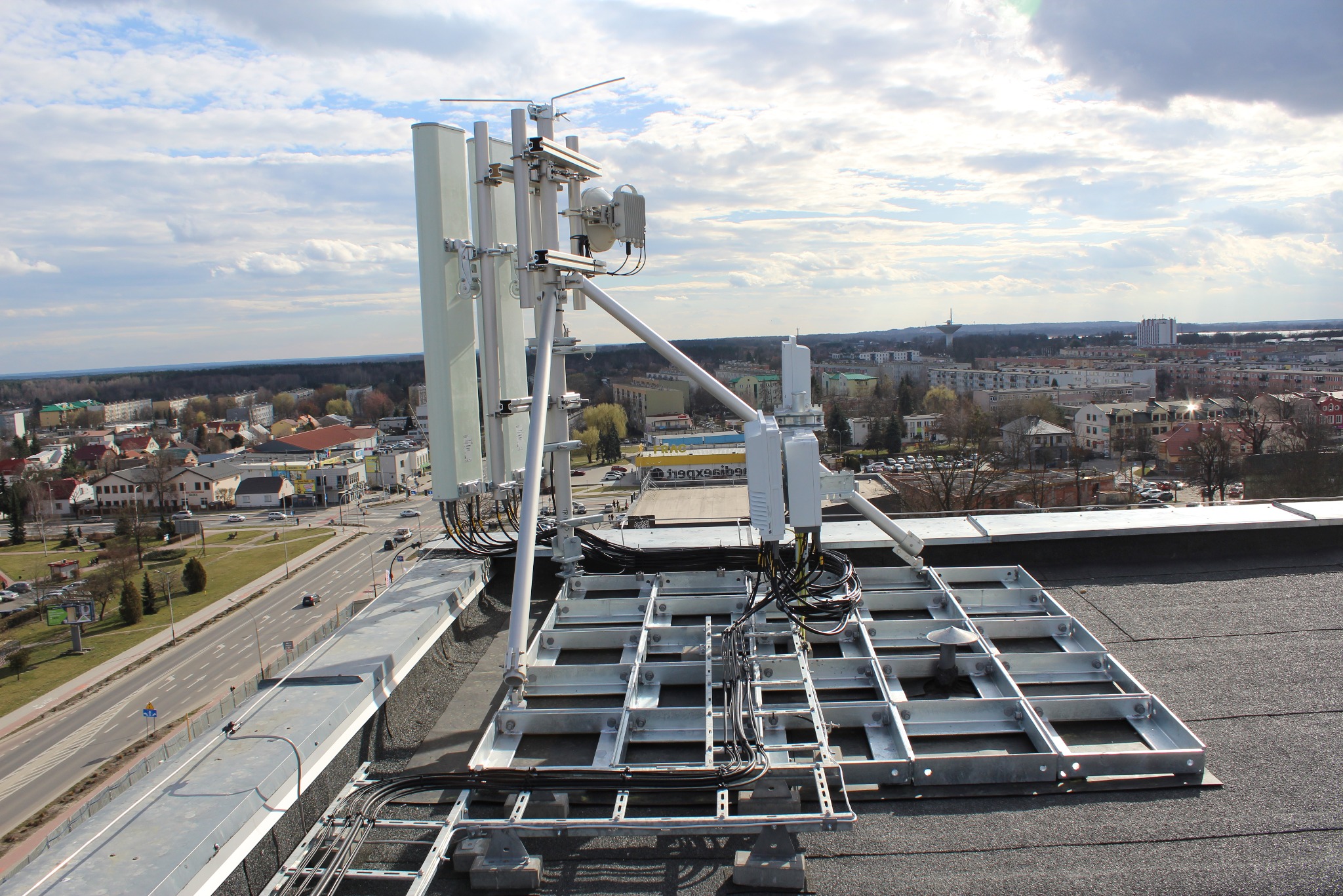 Anteny telekomunikacyjne zamontowane na dachu budynku z widokiem na miasto w tle. Metalowa konstrukcja wsporcza, kable i urządzenia. Słoneczny dzień.