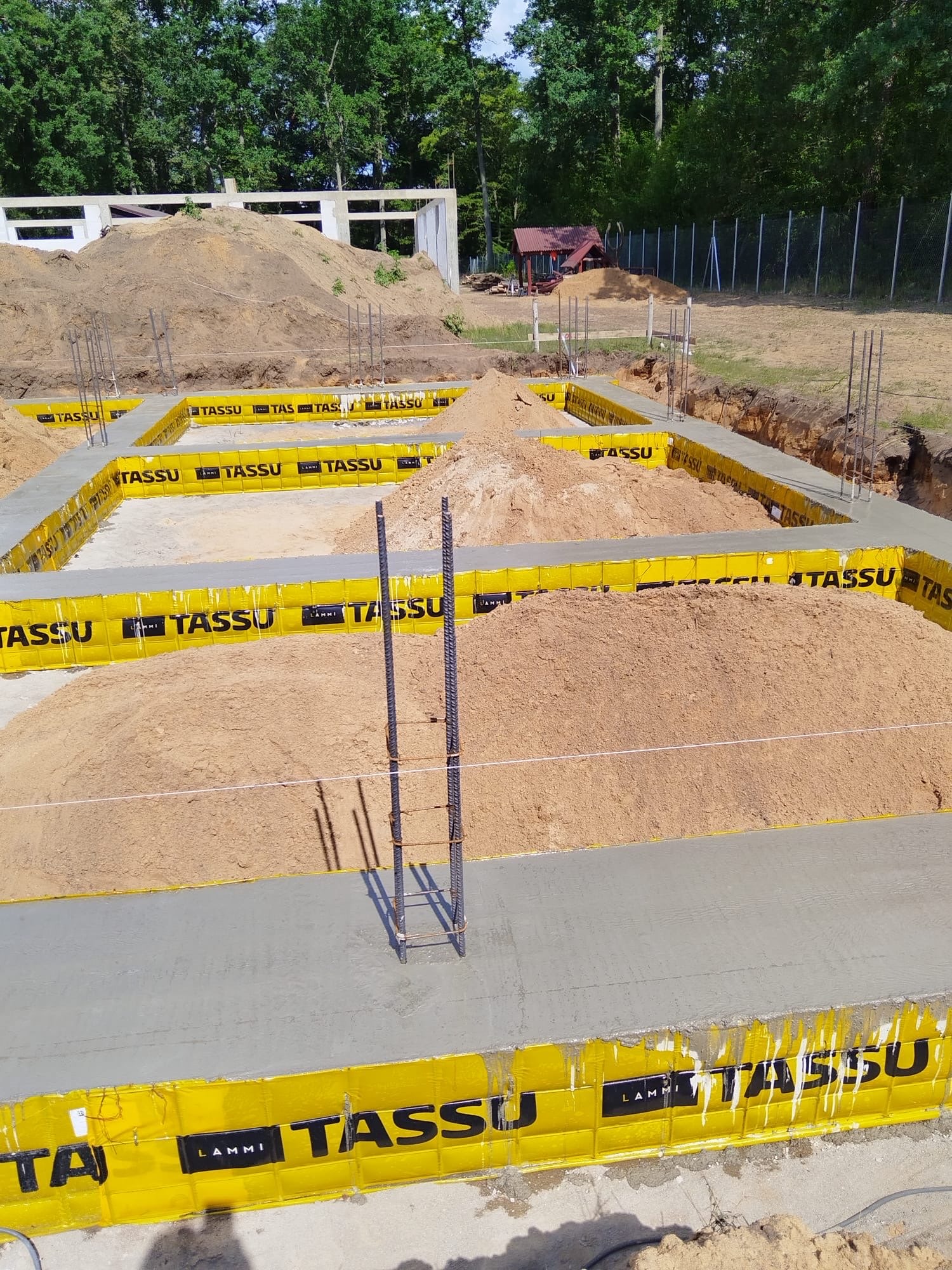 Zalane fundamenty z żółtym szalunkiem TASSU, wystające pręty zbrojeniowe, pryzmy piasku w wykopie. W tle szkielet budynku w stanie surowym otwartym i zieleń.