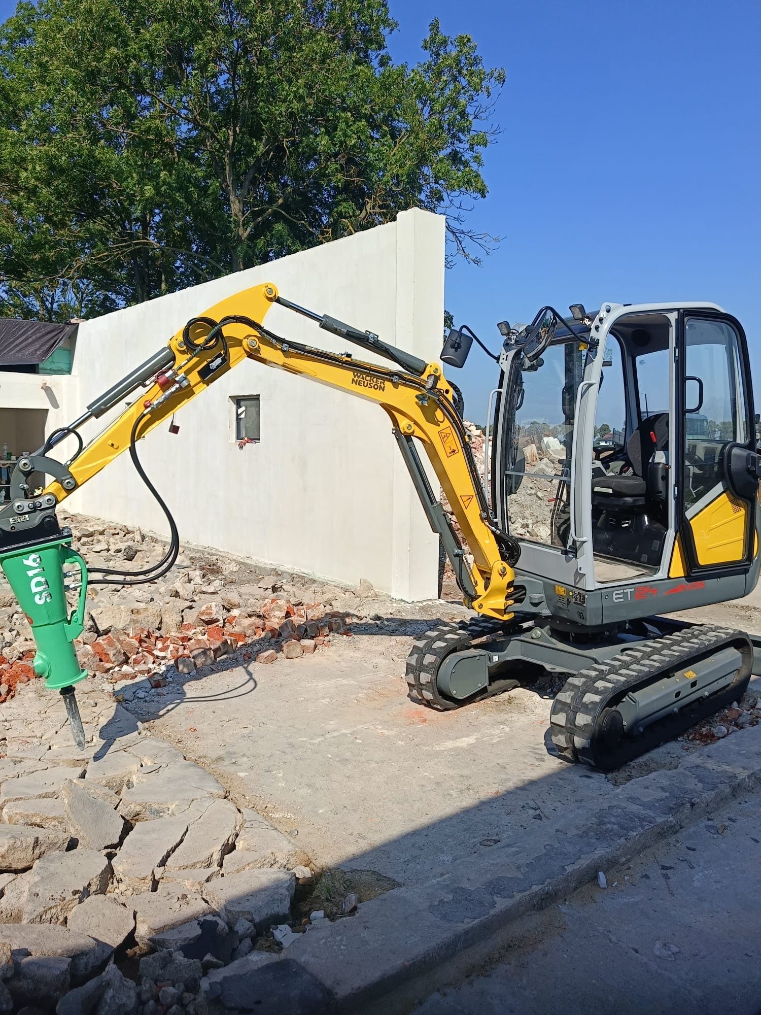Żółto-szara mini koparka Wacker Neuson z młotem pneumatycznym na gruzowisku, na tle białej ściany i błękitnego nieba. Widoczne ślady prac rozbiórkowych.