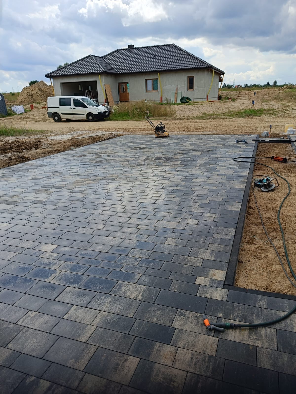 Częściowo ułożona, szara kostka brukowa na posesji w Chełmży. W tle dom w stanie surowym i biały samochód dostawczy. Widoczne narzędzia i węże.