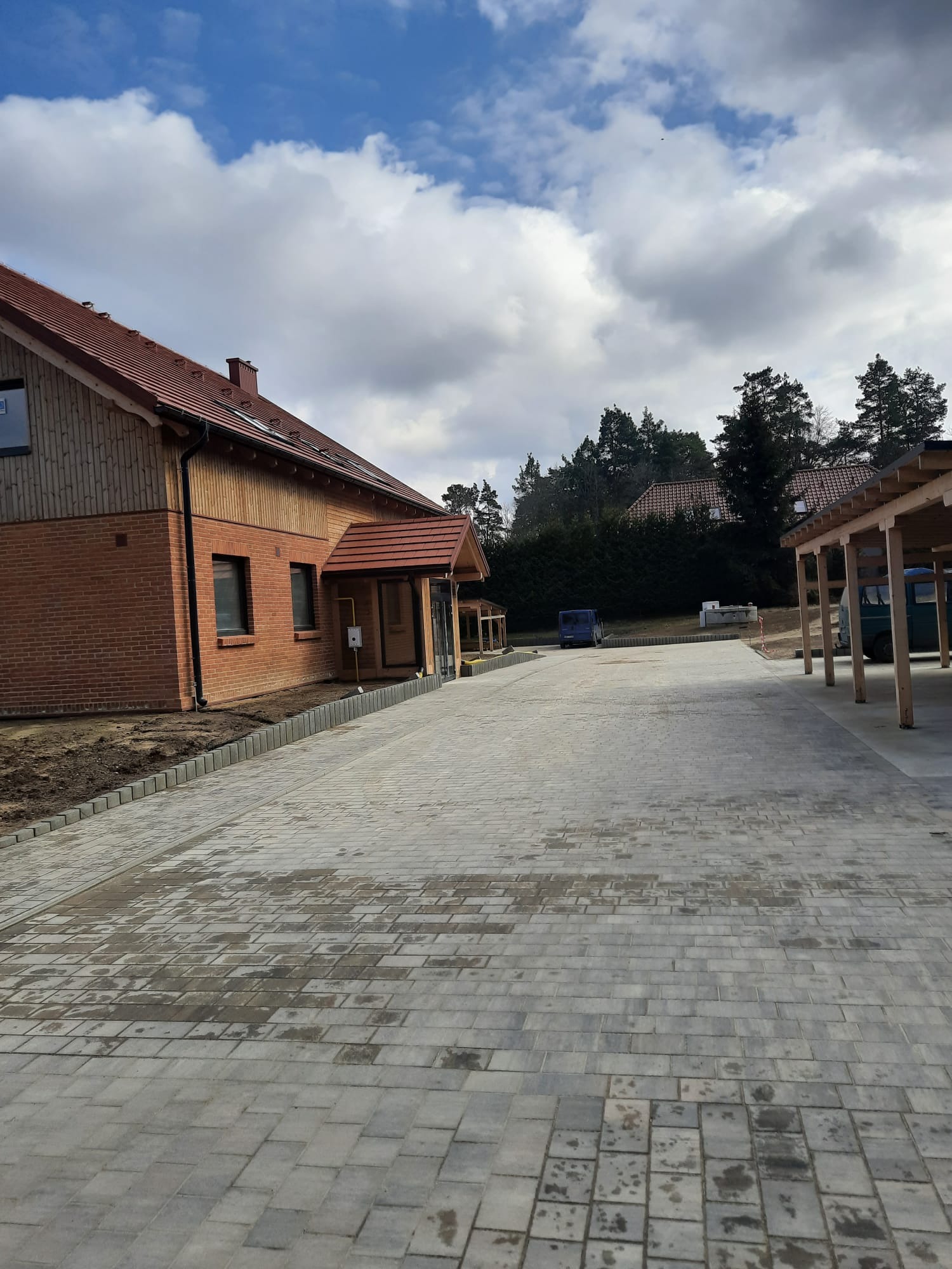 Ułożona, szara kostka brukowa prowadząca do domu z cegły i drewnianą wiatą garażową. Widoczne obrzeża i przygotowany teren pod nasadzenia. Pochmurne niebo w tle.