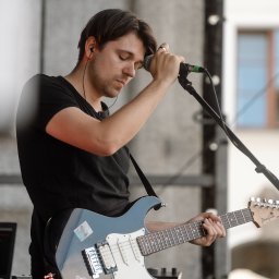 kostemusic - Gitarzysta z zamkniętymi oczami, w czarnej koszulce, grający na niebieskiej gitarze elektrycznej podczas występu na scenie z mikrofonem i laptopem w tle.