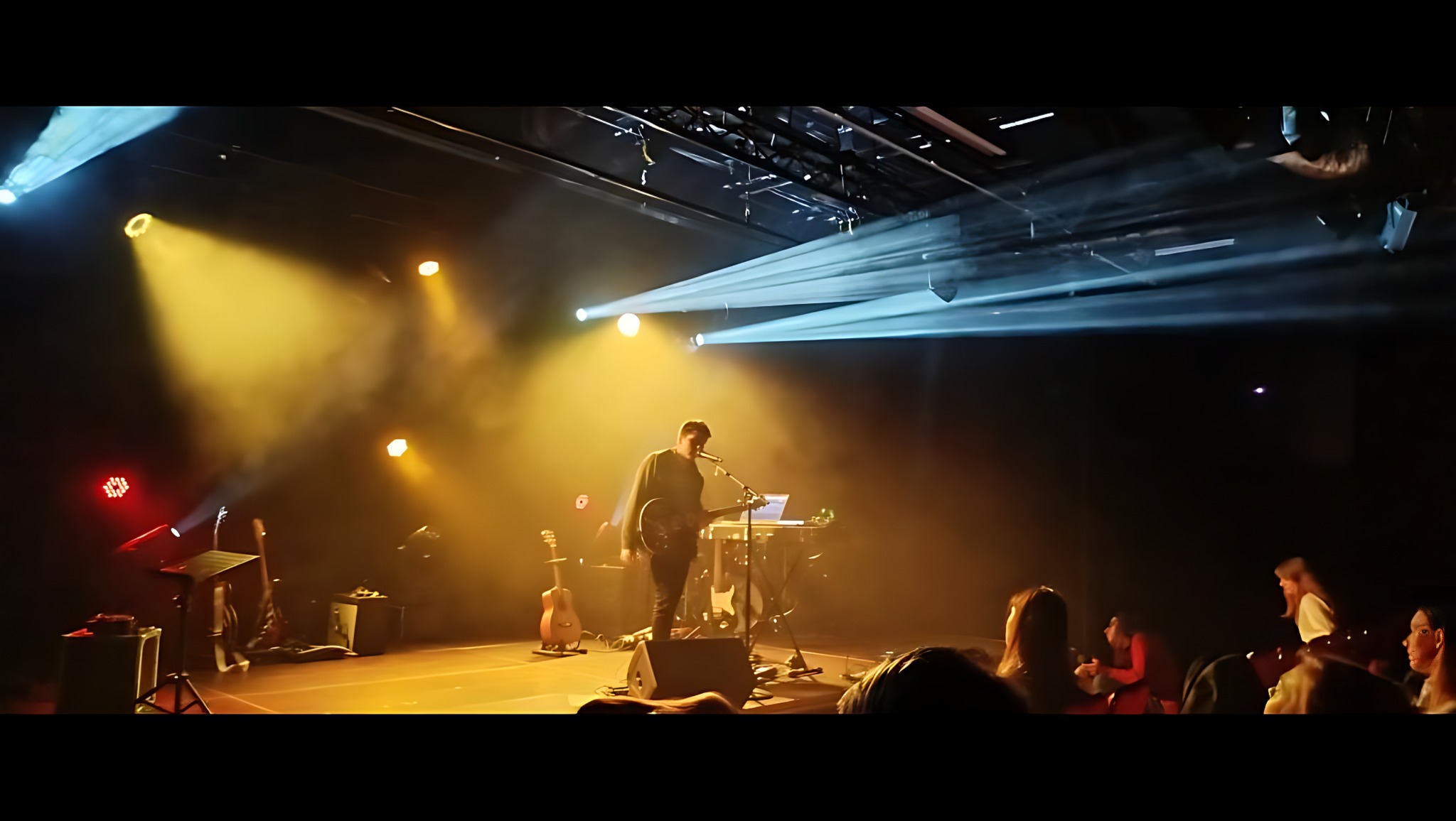 Wokalista z gitarą na scenie w oświetleniu reflektorów, z widoczną publicznością w tle. Scena z instrumentami i sprzętem nagłośnieniowym.