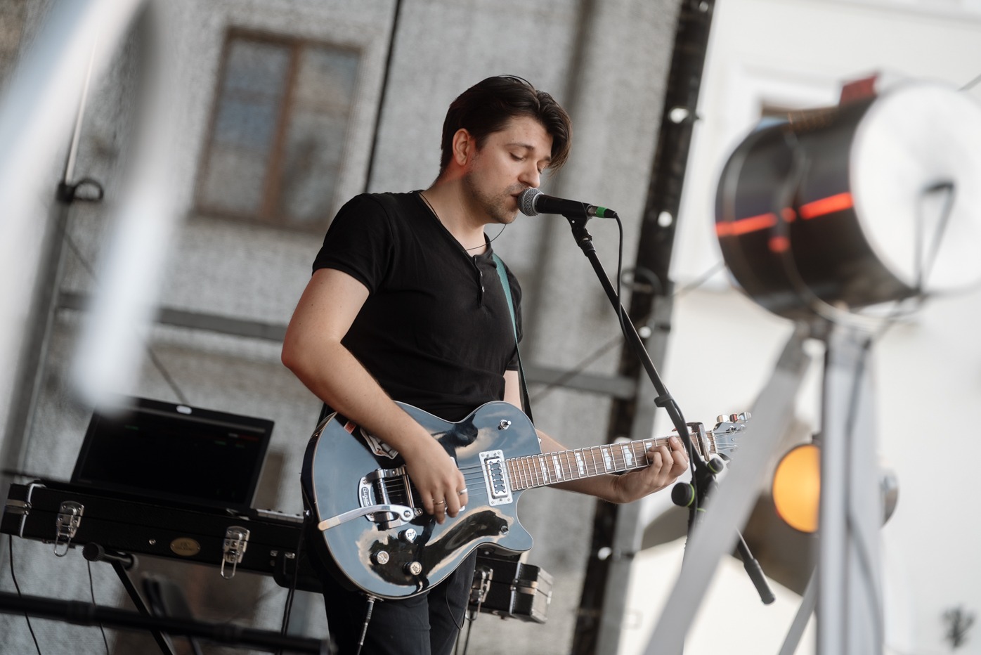 Gitarzysta w czarnym stroju śpiewa do mikrofonu, grając na elektrycznej gitarze na scenie z laptopem i perkusją w tle. Koncert na żywo.