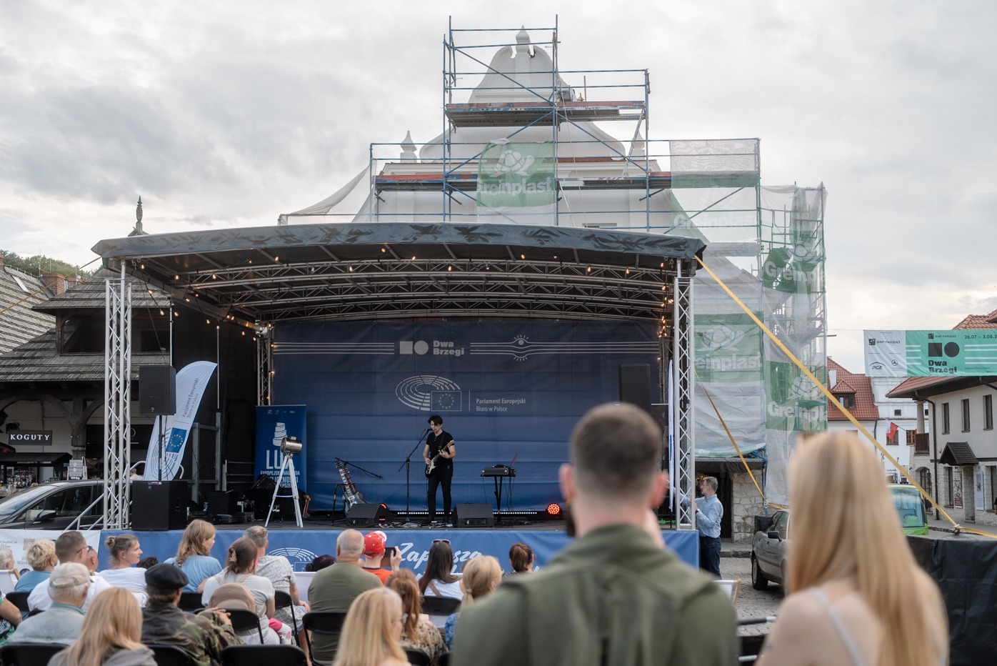 Koncert plenerowy: gitarzysta na scenie przed publicznością, w tle budynek w rusztowaniach i baner festiwalu 'Dwa Brzegi'.