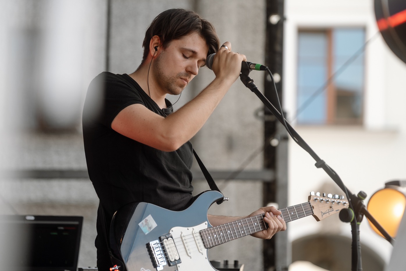 Gitarzysta z zamkniętymi oczami, w czarnej koszulce, grający na niebieskiej gitarze elektrycznej podczas występu na scenie z mikrofonem i laptopem w tle.