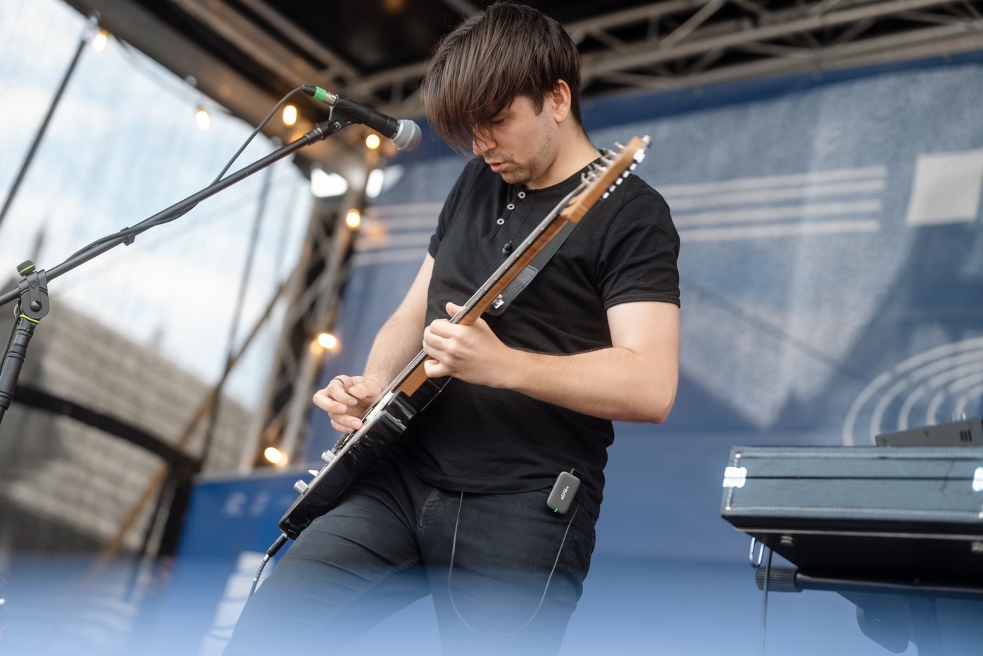 Gitarzysta w czarnym stroju gra na gitarze elektrycznej na scenie z mikrofonem i klawiszami w tle, oświetlony światłami.