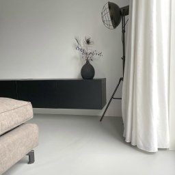 Mikrocem - Minimalistyczne wnętrze z jasną posadzką, czarną szafką RTV, lampą studyjną i zasłoną. Wazon z piórami pawimi dodaje elegancji.