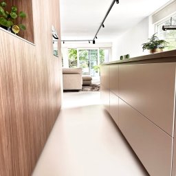 Mikrocem - Jasna, gładka posadzka w nowoczesnym wnętrzu z drewnianymi elementami zabudowy i oświetleniem szynowym. Widok na salon z sofą przez otwarte przejście.