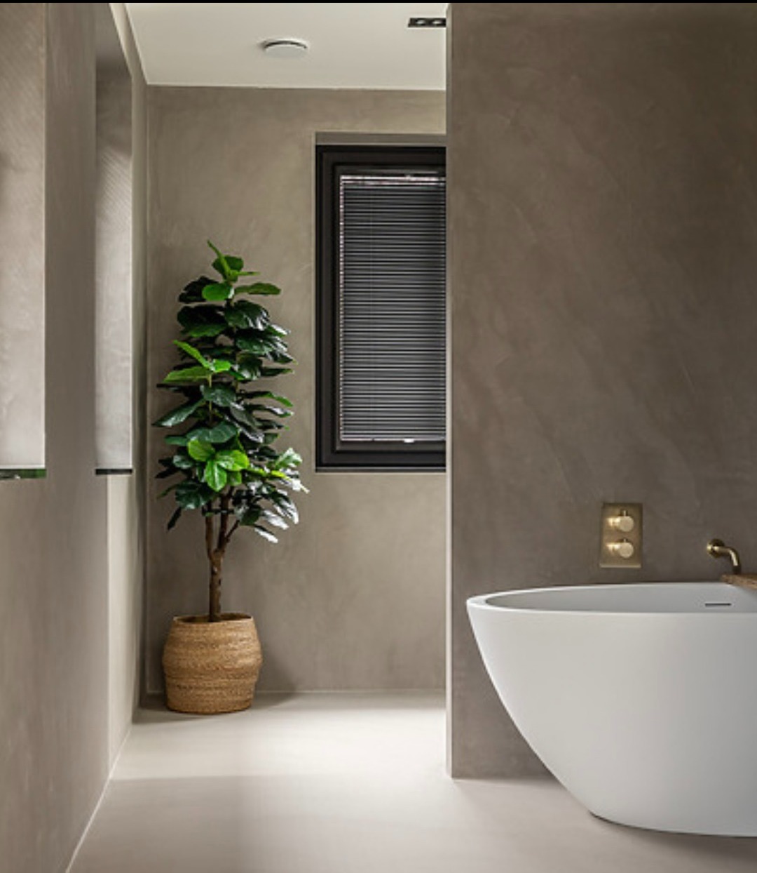 Stylowa łazienka z mikrocementem: gładkie, szare ściany i podłoga, biała wanna wolnostojąca, roślina w plecionym koszu, czarne okno z żaluzjami. Minimalistyczny design.