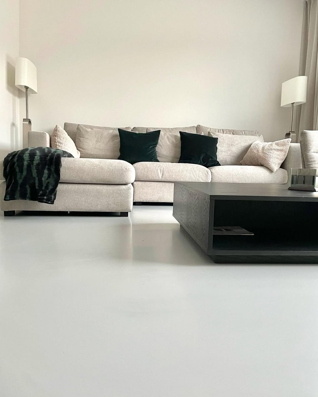 Jasnoszara, gładka posadzka w salonie z beżową sofą i ciemnym stolikiem kawowym. Minimalistyczne wnętrze z lampami podłogowymi i zasłonami w tle.