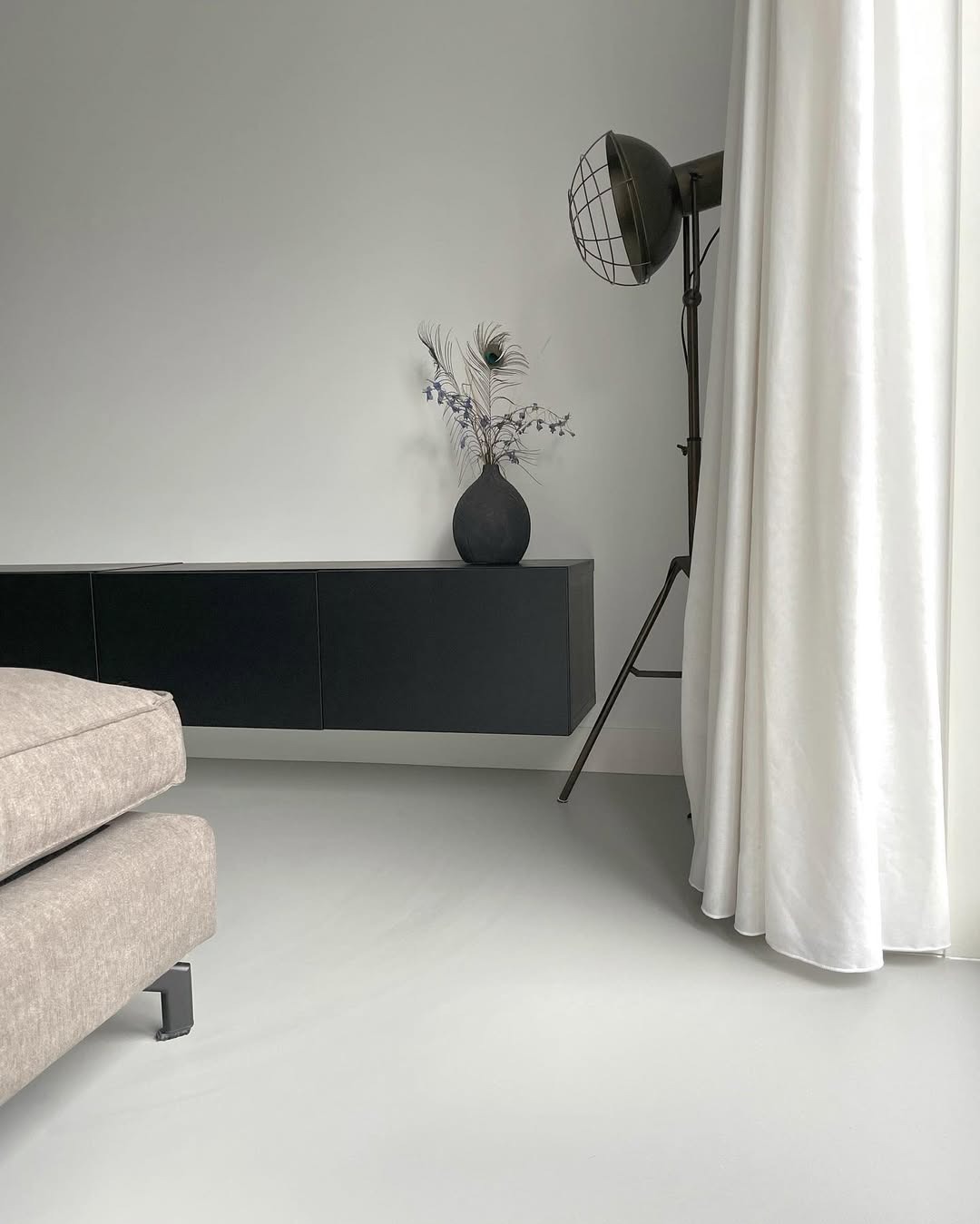 Minimalistyczne wnętrze z jasną posadzką, czarną szafką RTV, lampą studyjną i zasłoną. Wazon z piórami pawimi dodaje elegancji.