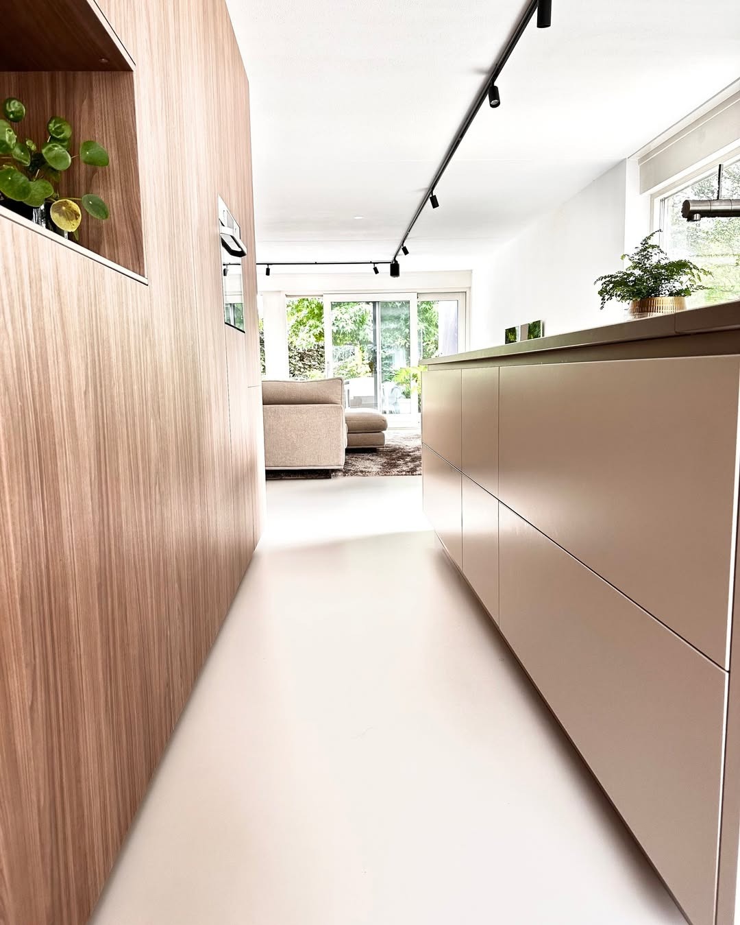 Jasna, gładka posadzka w nowoczesnym wnętrzu z drewnianymi elementami zabudowy i oświetleniem szynowym. Widok na salon z sofą przez otwarte przejście.