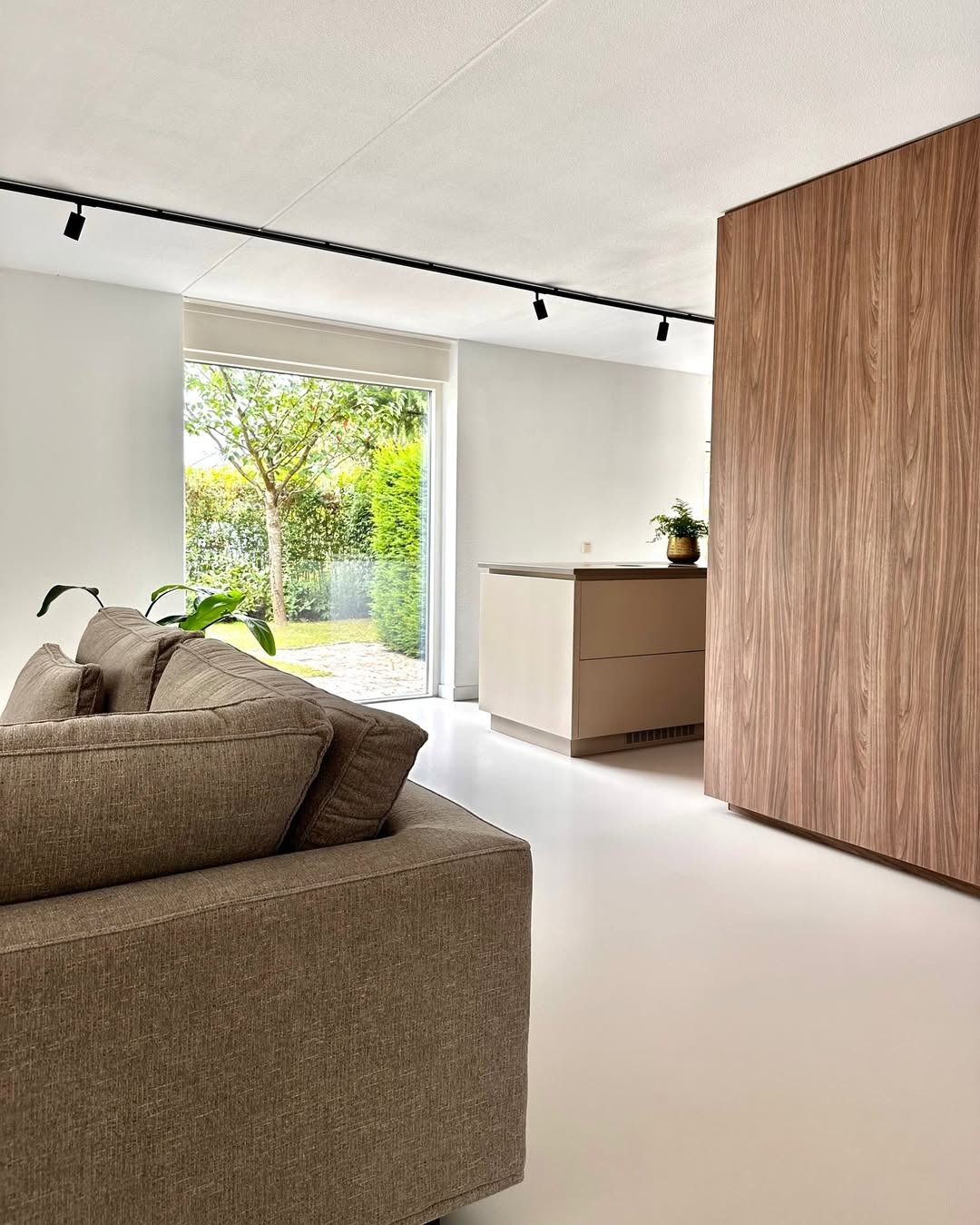 Jasne wnętrze z beżową sofą, białymi ścianami i podłogą. Widoczne okno z widokiem na zieleń, drewniana zabudowa i minimalistyczne oświetlenie szynowe.