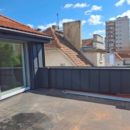 Ustrzel Dach Kamil Ciesielski - Dach pokryty papą z obróbką blacharską w kolorze grafitowym, widok na budynki w tle i błękitne niebo. Na dachu leżą rury.