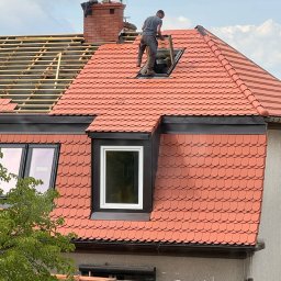 Ustrzel Dach Kamil Ciesielski - Czerwony dach z dachówką karpiówką w trakcie montażu okna dachowego. Widoczna więźba dachowa i komin z cegły. Praca na wysokości.