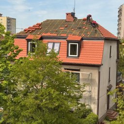 Ustrzel Dach Kamil Ciesielski - Dwóch dekarzy pracuje na dachu domu z czerwonej dachówki, układając nowe pokrycie. Widoczne rusztowania i zieleń drzew. Ujęcie z góry, w tle budynki mieszkalne.