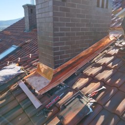 Ustrzel Dach Kamil Ciesielski - Montaż obróbki blacharskiej komina z miedzianej blachy na dachu z dachówki ceramicznej. Widoczne narzędzia i fragment sylwetki dekarza. Słoneczny dzień.