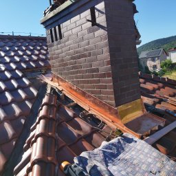 Ustrzel Dach Kamil Ciesielski - Ceglany komin z miedzianą obróbką blacharską na tle brązowej dachówki i błękitnego nieba. Widoczne narzędzia i fragment płachty zabezpieczającej.