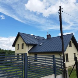 Ustrzel Dach Kamil Ciesielski - Dom jednorodzinny z ciemnoszarym dachem z blachy na tle błękitnego nieba z chmurami. Widoczny fragment ogrodzenia i słup elektryczny.