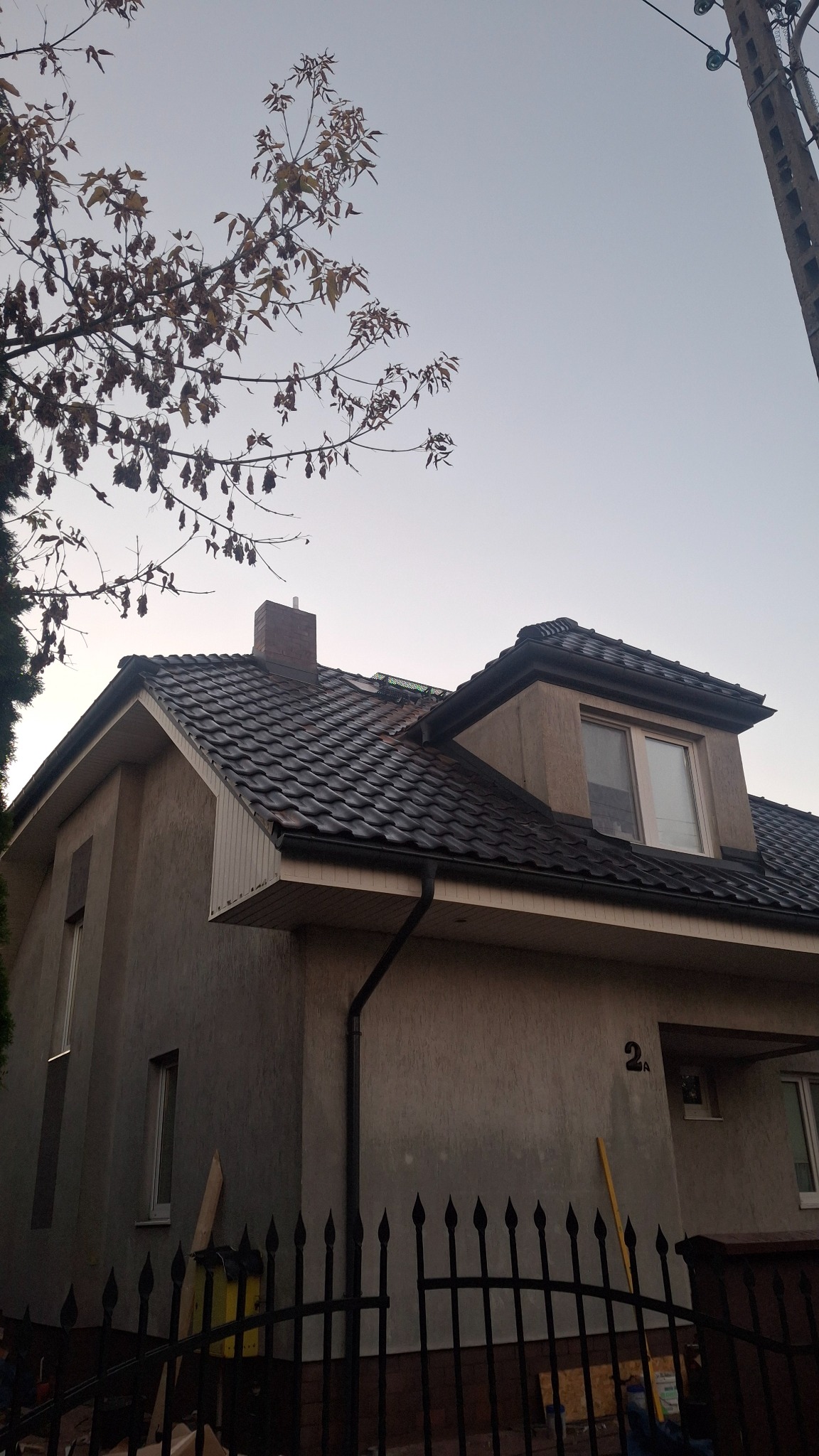 Dwukondygnacyjny dom z ciemną dachówką karpiówką i czarnymi rynnami, widziany zza metalowego ogrodzenia. Na elewacji numer domu '2A'.
