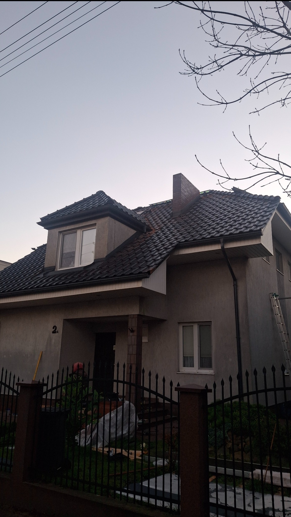 Dom z ciemną dachówką karpiówką i ceglanym kominem, widoczny zza metalowego ogrodzenia. Częściowo widoczna drabina oparta o ścianę budynku.