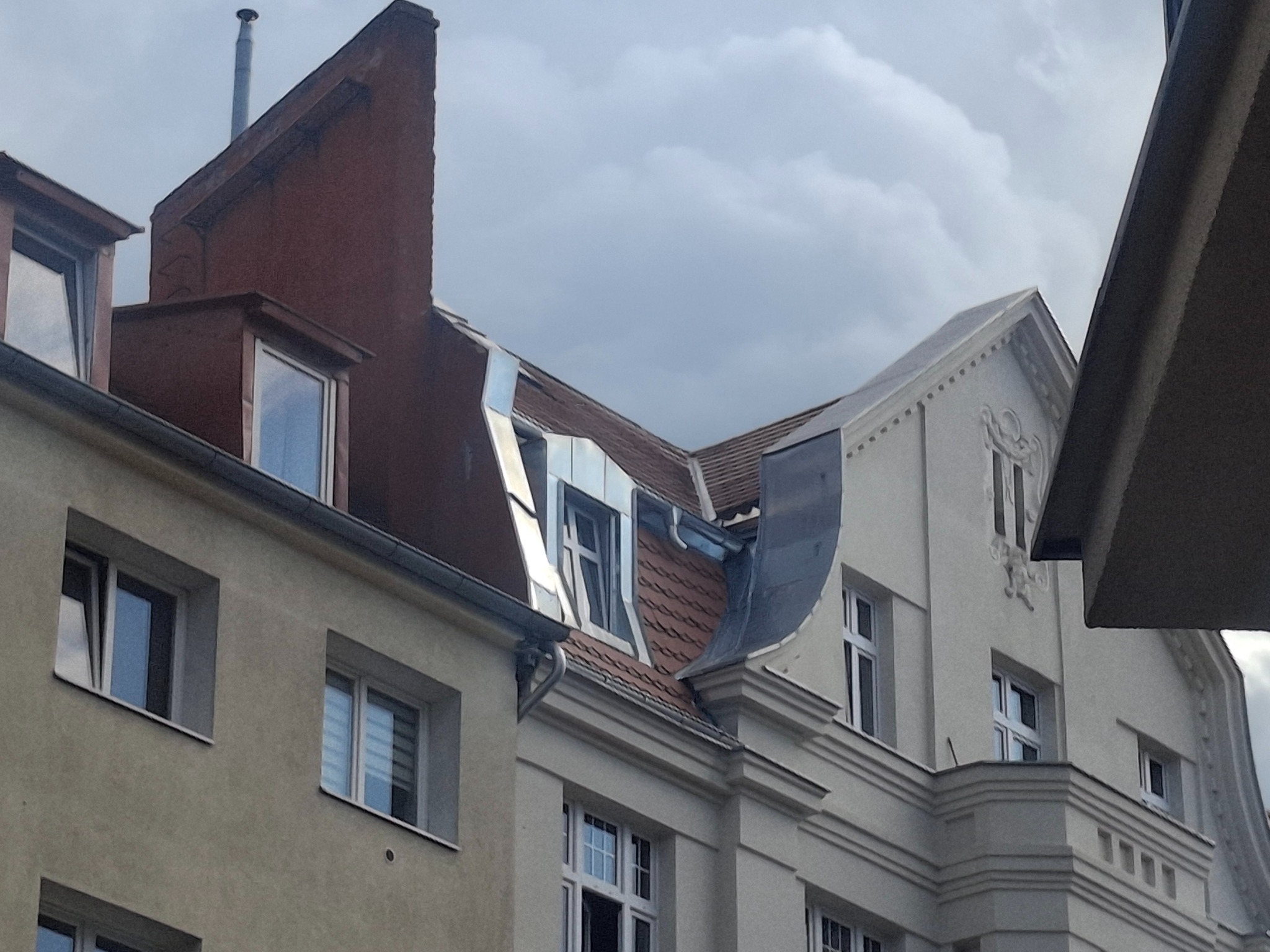 Fragmenty dachów kamienic z widocznymi oknami dachowymi, obróbkami blacharskimi i rynnami, na tle pochmurnego nieba. Ujęcie z dołu.